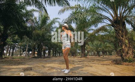 Woman tourist with plait walks looking around at growing young trees with lush leaves at oil palm farm elaeis guineensis on sunny day. Stock Photo