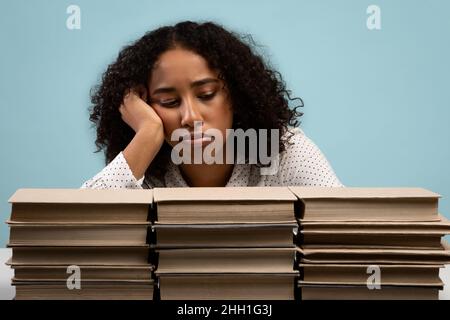 Exhausted young Adrican American woman sitting with big stacks of books, preparing for exam, feeling bored or tired Stock Photo