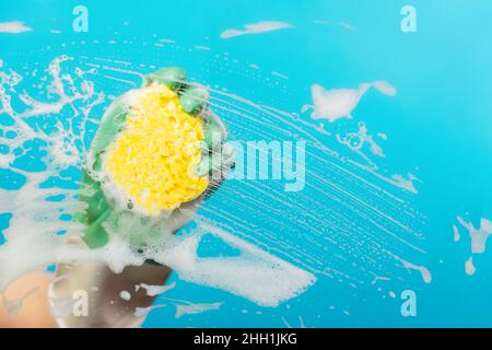 Close up of a hand in rubber gloves wipes the glass with a yellow foam sponge. Blue background with copy space. The concept of professional cleaning s Stock Photo