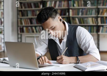 Focused serious Asian student guy with wireless earphones writing notes Stock Photo
