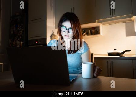 Caucasian woman wearing glasses at a wireless computer at night in the kitchen. Girl works overtime in the home office. Freelancer is typing on a Stock Photo