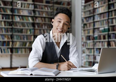 Happy Asian student with wireless earphones studying in library headshot Stock Photo