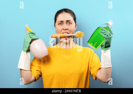 Portrait of a stressed Caucasian young woman in rubber gloves, holding a brush in her mouth and household cleaning products in her hands. Blue backgro Stock Photo