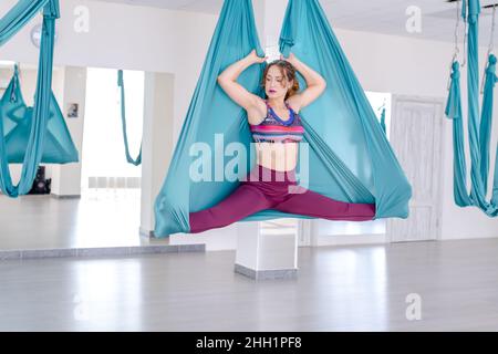 attractive young woman doing antigravity yoga using blue hammock in studio. Lady doing side splits. Stock Photo