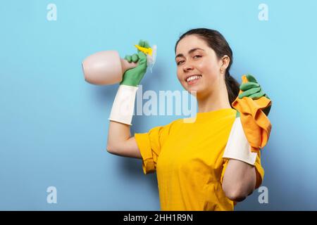 Portrait of a smiling Caucasian young woman in rubber gloves, holding a sprayer and rag in her hands. Blue background with copy space. Concept of clea Stock Photo