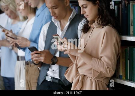 Diverse group of digital addicted students standing at bookshelves Stock Photo
