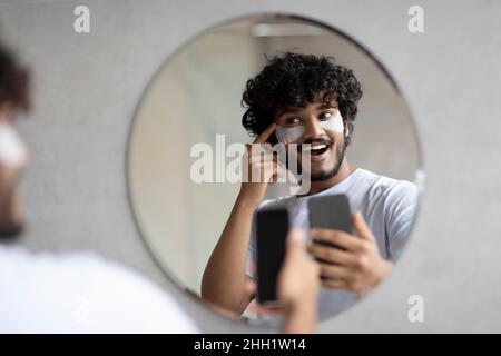 Excited indian man with eye patches taking photo on cellphone, looking and smiling at mirror in bathroom interior Stock Photo