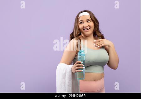 Cheerful pretty caucasian plus size young woman in sport wear holding towel and water bottle look at free space Stock Photo