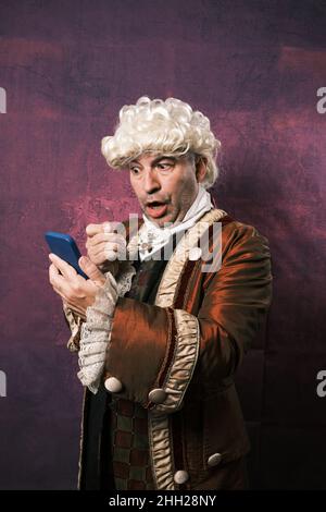 Man in classical renaissance clothes with a surprised expression on his face while looking at the mobile phone screen with a monocle. Stock Photo