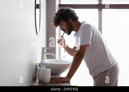 Healthy teeth concept. Profile side view of indian bearded guy brushing teeth with paste, standing near sink in bathroom Stock Photo