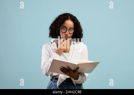 Pensive black female student in glasses reading textbook, thinking over assignment, studying for exam on blue background Stock Photo