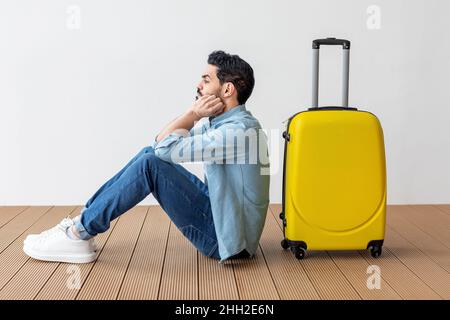 Upset arab male tourist sitting near suitcase and looking away at copy space, waiting for flight over light wall Stock Photo