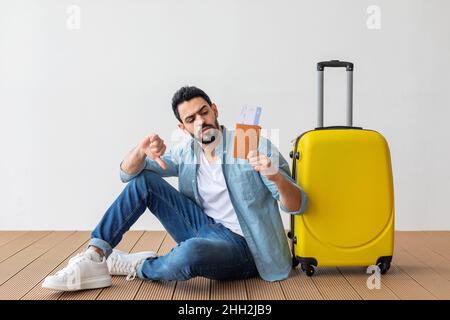 Bad travel agency service. Young arab man holding passport with tickets, showing dislike gesture, sitting near suitcase Stock Photo