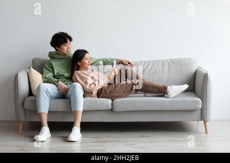 Affectionate young Asian couple using smartphone, relaxing on couch together at home, free space Stock Photo