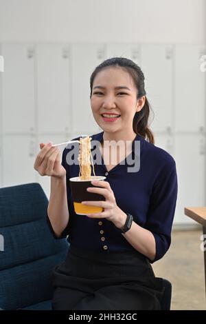 Smiling asian woman holding plastic fork eating instant noodle in office. Stock Photo