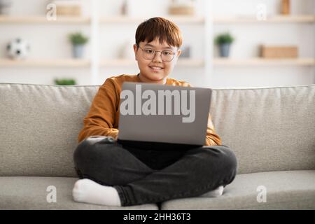 Happy chinese kid sitting on couch with laptop at home Stock Photo