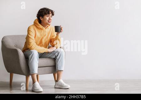 Handsome young Asian man drinking coffee, having break, chilling in armchair with hot drink against white studio wall Stock Photo