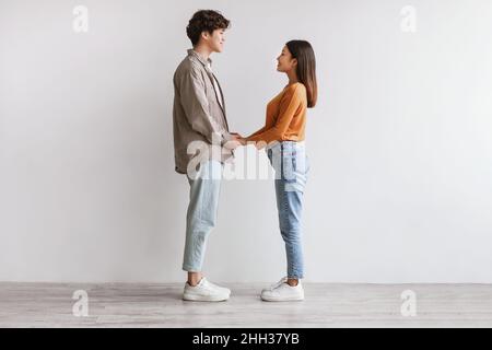 Full length of millennial Asian couple holding hands and looking at each other against white studio wall, side view Stock Photo