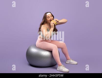 Happy funny pretty hungry caucasian plus size young lady in sports uniform sits on fit ball and eating burger