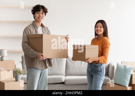Cheerful young Asian woman and her husband holding carton boxes, smiling at camera in new home. Moving day concept Stock Photo