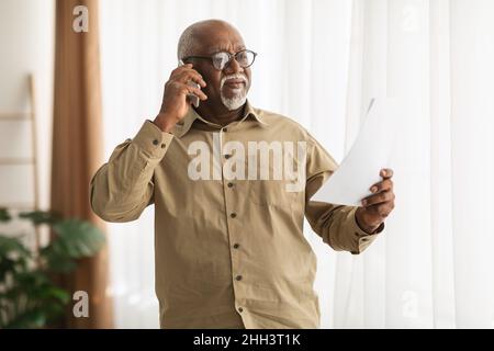 Mature Black Man Talking On Cellphone Holding Paper Standing Indoors Stock Photo