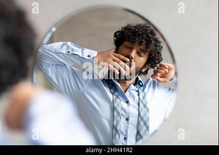 Sleepy indian businessman yawning while standing in bathroom and looking at his reflection in mirror Stock Photo