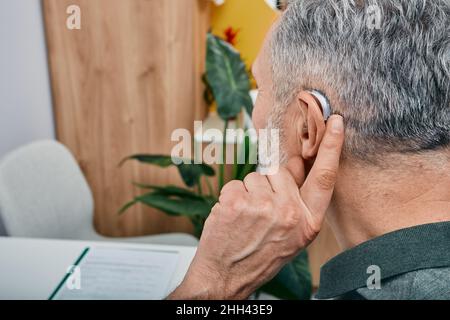 Hearing solutions for elderly deafness people. Older gray-haired man tunes his hearing aid behind the ear by pressing his finger on setting button whi Stock Photo