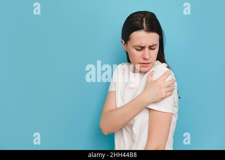 Unhappy sad tired young brunette woman has painful feelings in left shoulder, clenches teeth, wears white t-shirt, posing isolated on blue studio back Stock Photo