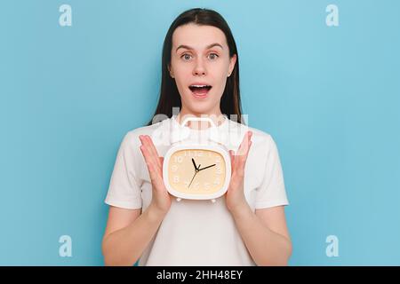 Portrait of surprised amazed young caucasian woman 20s old years wearing casual white t-shirt holding in hand clock, shocked looking camera, posing is Stock Photo