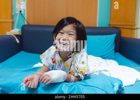 Illness asian child smiling happily and looking at camera. Girl admitted in hospital while saline intravenous (IV) on hand. Health care stories. Stock Photo