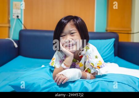 Illness asian child smiling happily and looking at camera. Girl admitted in hospital while saline intravenous (IV) on hand and propping up her face. H Stock Photo