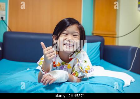 Illness asian child smiling happily and showing thumb up hand sign. Girl admitted in hospital while saline intravenous (IV) on hand. Health care stori Stock Photo