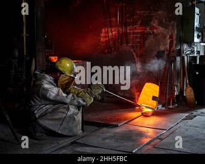 Craftsperson wearing protective workwear pouring cast metal in steel mill Stock Photo