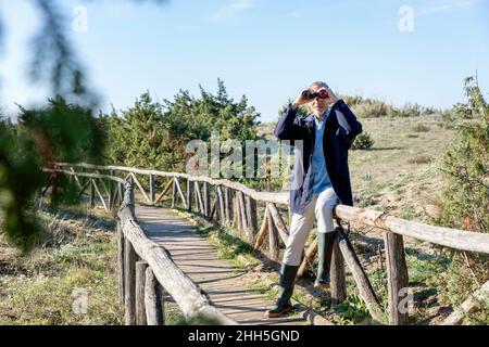 Man looking through binoculars sitting on railing at wooden bridge Stock Photo