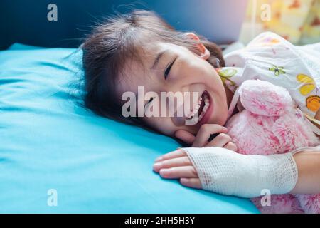 Close up. Illness asian child smiling happily and looking at camera. Girl admitted in hospital while saline intravenous (IV) on hand. Health care stor Stock Photo