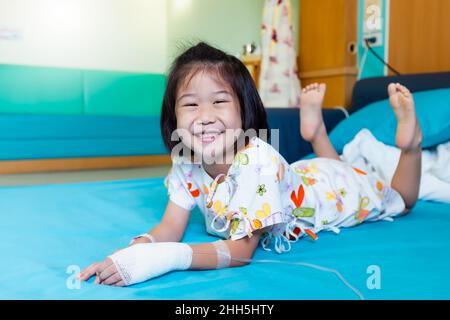 Illness asian child smiling happily and looking at camera. Girl admitted in hospital while saline intravenous (IV) on hand. Health care stories. Stock Photo
