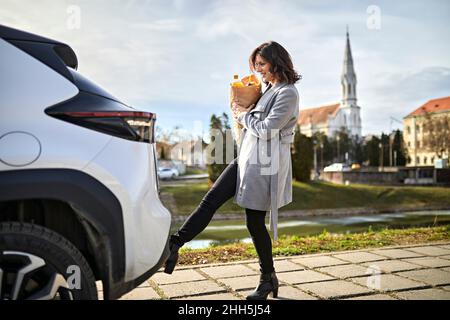 Woman carrying grocery bag opening car trunk with foot sensor Stock Photo