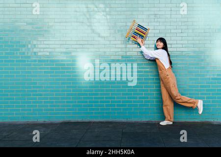 Smiling young woman holding abacus in front of turquoise brick wall Stock Photo