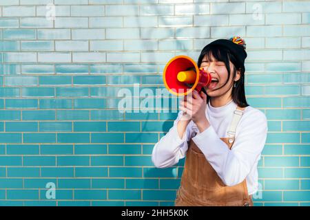 Woman screaming through megaphone in front of turquoise brick wall Stock Photo