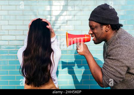 Angry man shouting through megaphone on woman with head in hands facing wall Stock Photo
