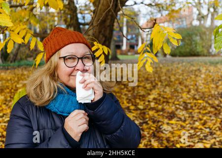 Senior woman blowing nose with tissue paper in autumn park Stock Photo