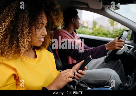Woman using smart phone and friend driving car on road trip Stock Photo