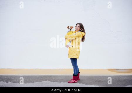 Smiling young woman wearing raincoat carrying dog on road Stock Photo