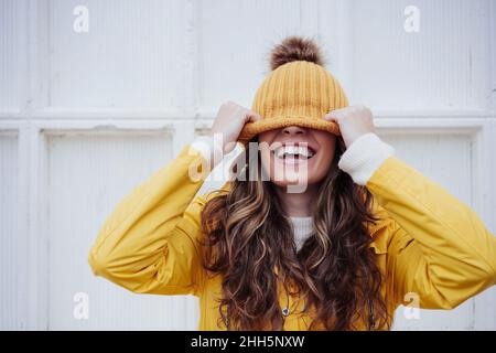 Playful woman covering face with knit hat in front of wall Stock Photo