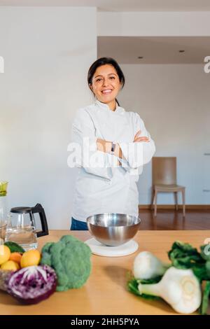 Confident chef with arms crossed standing by table in kitchen at home Stock Photo
