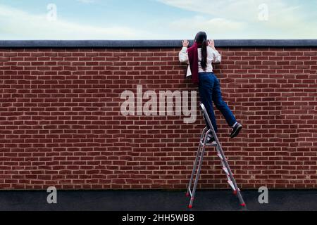 Young woman standing on ladder peeking over wall Stock Photo