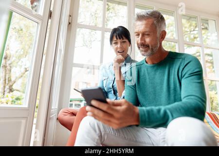 Man using smart phone sitting by surprised woman at home Stock Photo