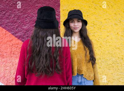 Smiling young woman standing by sister facing colorful wall Stock Photo