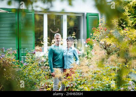 Smiling mature couple standing in backyard seen through plants Stock Photo