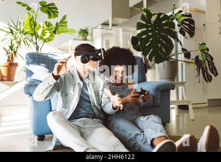 Young man with VR glasses gesturing by cheerful girlfriend playing video game at home Stock Photo